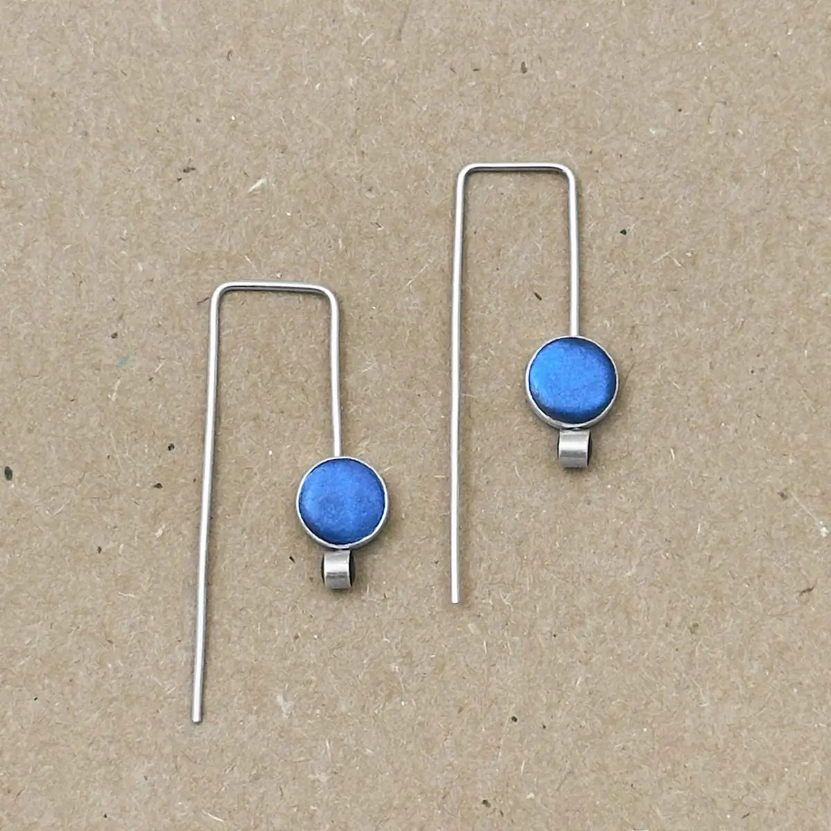 Flat lay of Orbit Drop Earrings in sterling silver with peacock blue hand-tinted clay circles on natural linen background.