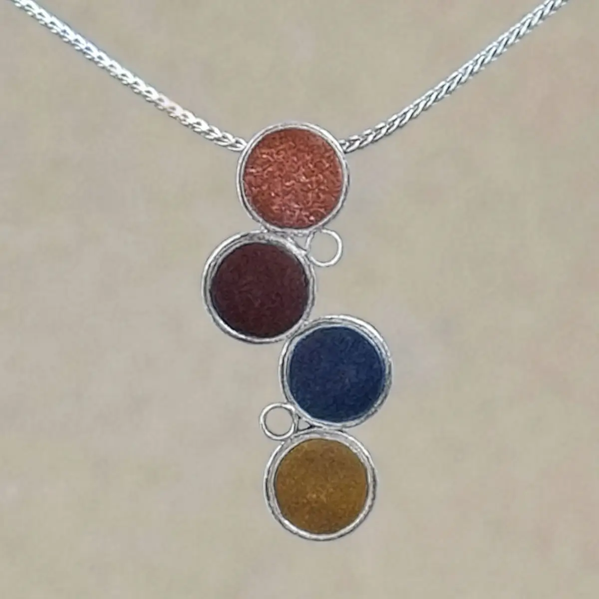 Close-up of the Cascade Pendant showing the metallic sheen and fine glitter texture of four hand-tinted clay discs in terracotta, burgundy, slate gray, and ochre, set in sterling silver circles.