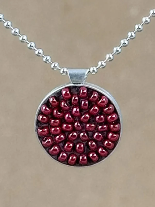Detail of round sterling silver pendant filled with brick-red seed beads set in warm brown clay, hung on a silver bead chain against a natural linen background.
