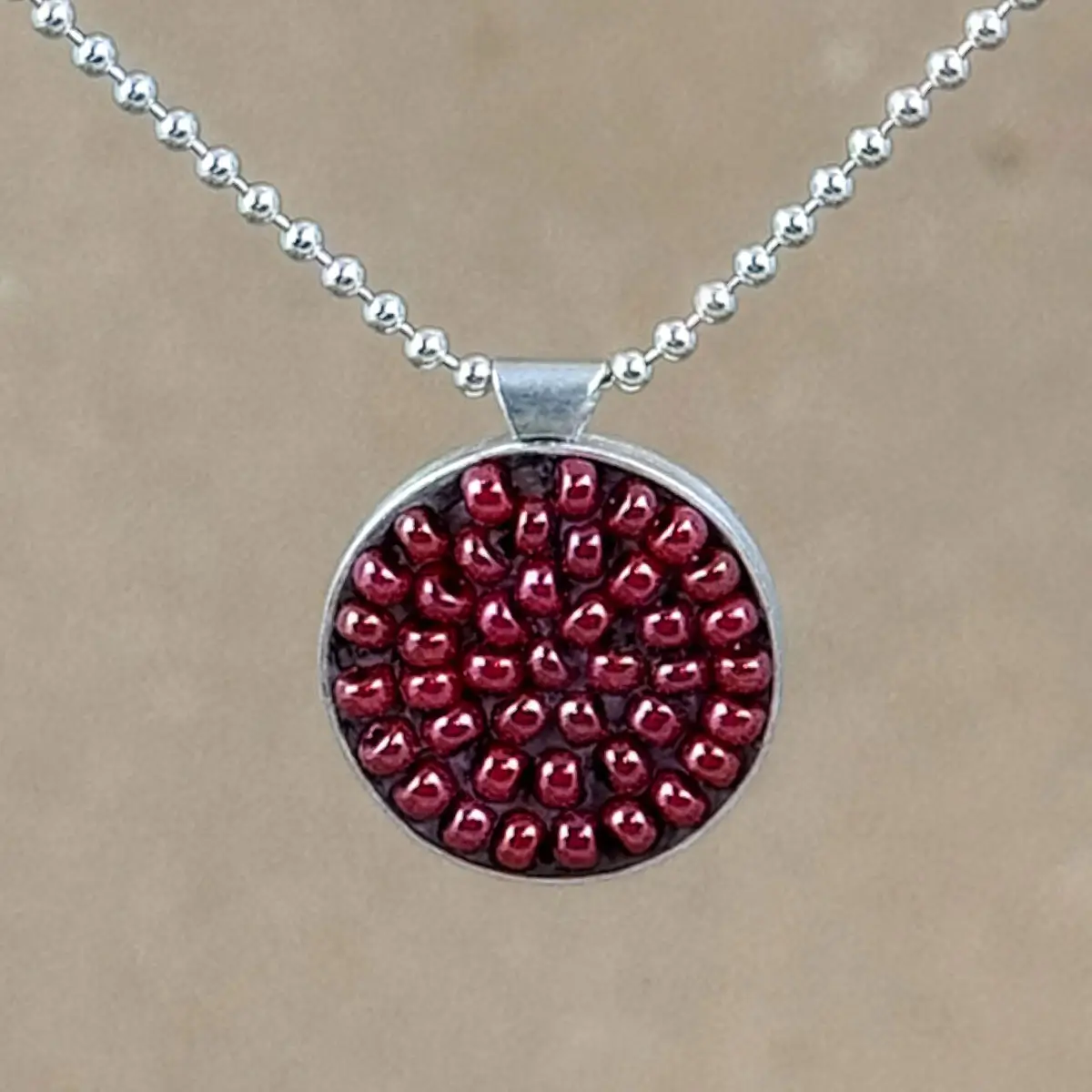 Detail of round sterling silver pendant filled with brick-red seed beads set in warm brown clay, hung on a silver bead chain against a natural linen background.