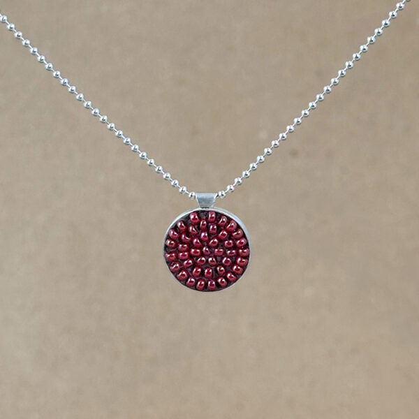Round sterling silver pendant filled with brick-red seed beads set in warm brown clay, hung on a silver bead chain against a natural linen background.