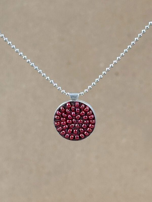 Round sterling silver pendant filled with brick-red seed beads set in warm brown clay, hung on a silver bead chain against a natural linen background.