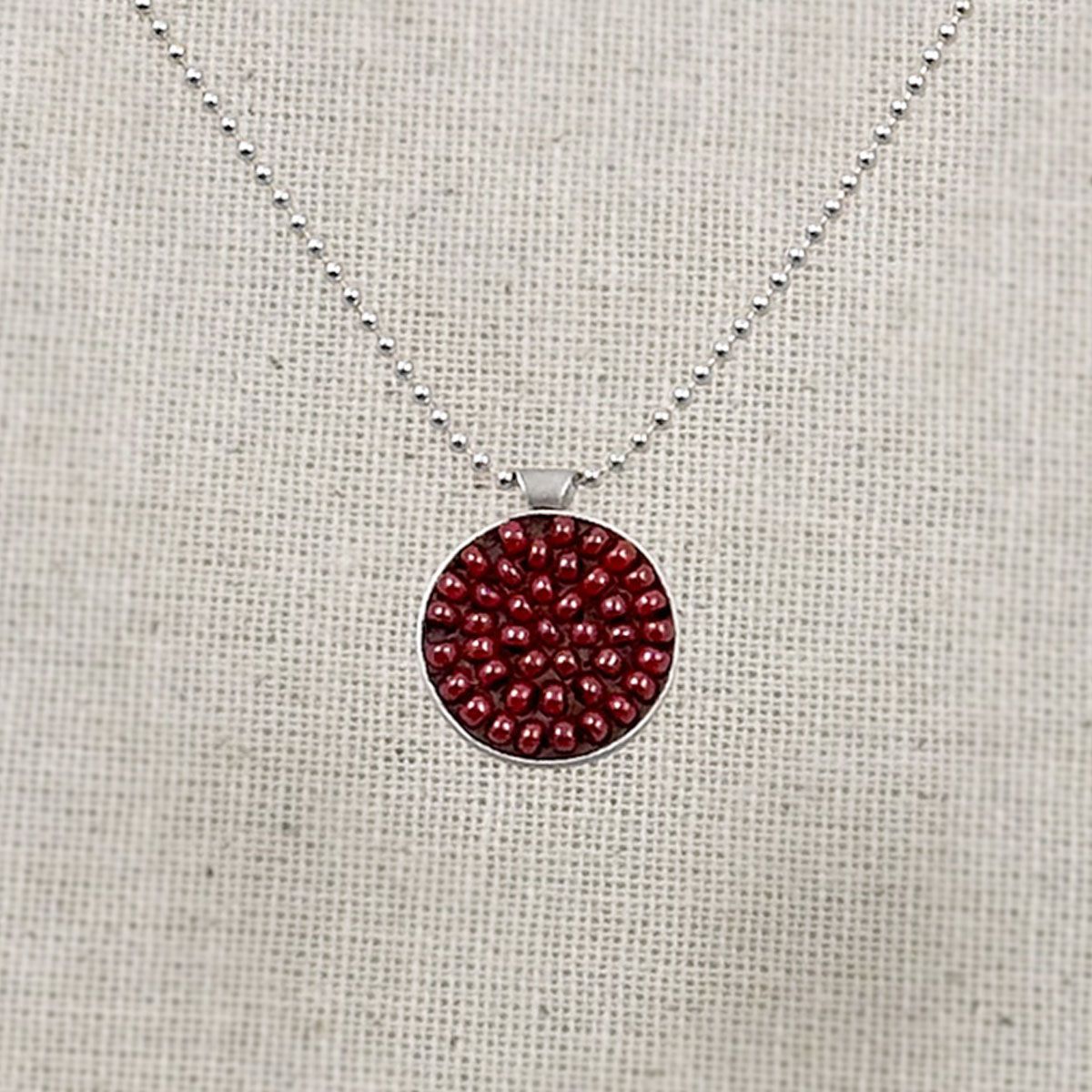 Round sterling silver pendant filled with brick-red seed beads set in warm brown clay, hung on a silver bead chain against a natural linen background.