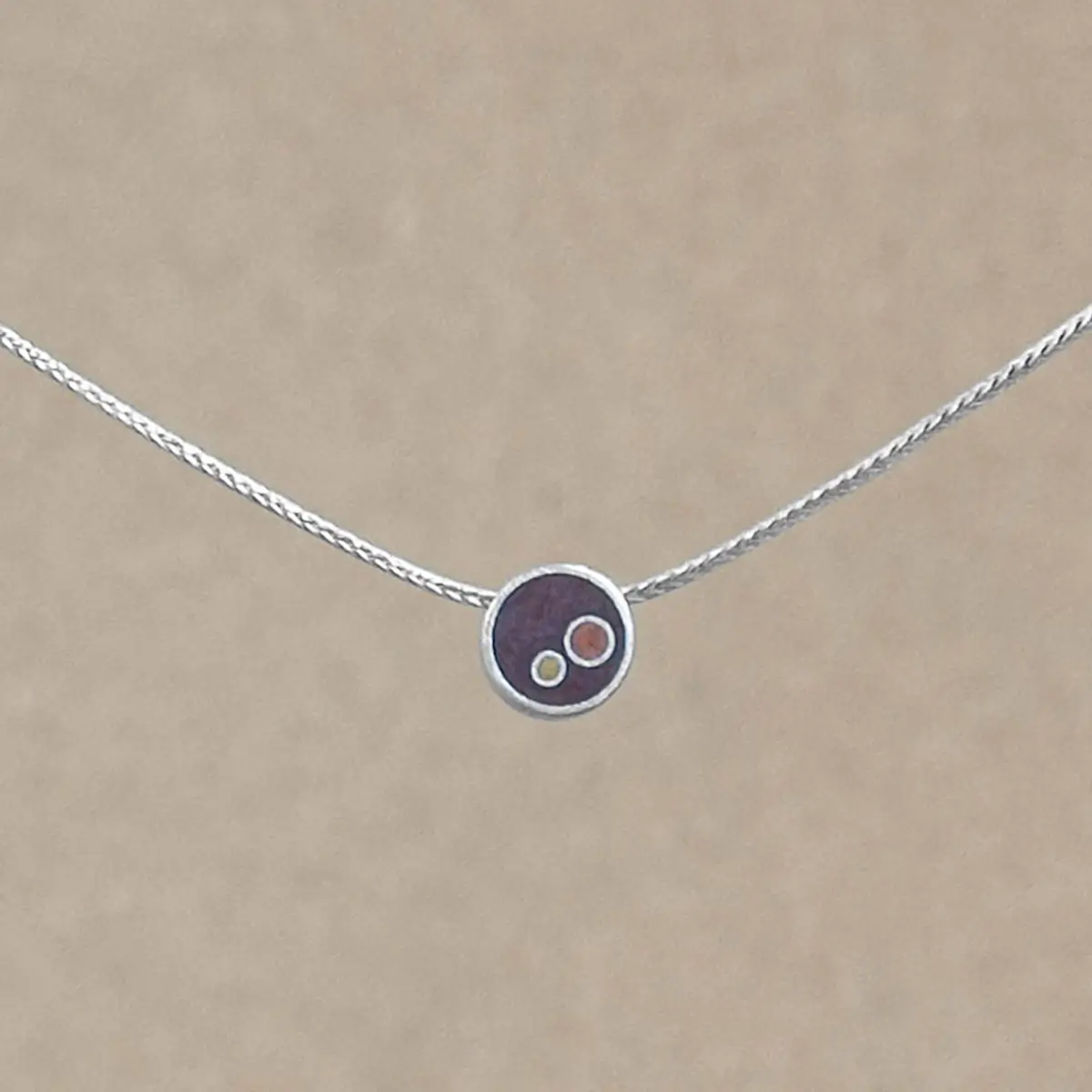 A small sterling silver circle pendant on a silver foxtail chain, centered against a warm beige background. The pendant holds deep brownish-copper hand-tinted clay inset with two smaller circles in gold and copper tint.