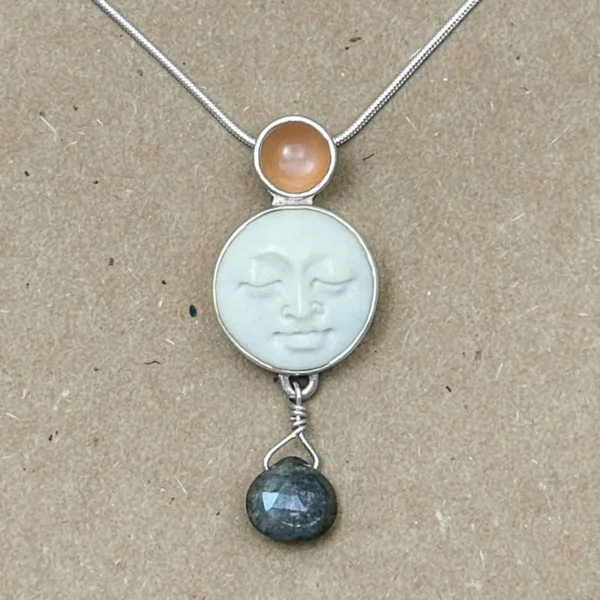 Close-up of a sterling silver pendant with a hand-carved bone moon face cabochon, small round peach moonstone set above, and a dark faceted labradorite briolette suspended below on a wire-wrapped loop.