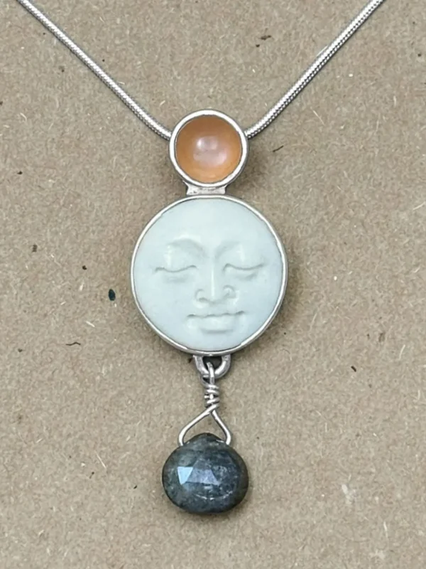 Close-up of a sterling silver pendant with a hand-carved bone moon face cabochon, small round peach moonstone set above, and a dark faceted labradorite briolette suspended below on a wire-wrapped loop.