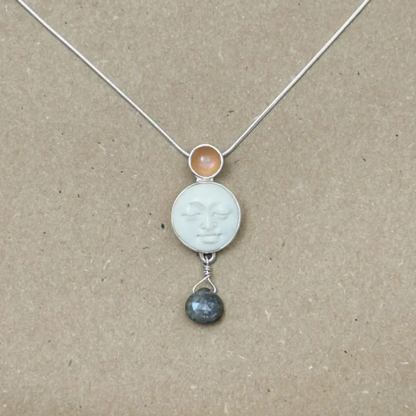Sterling silver pendant necklace featuring a hand-carved bone moon face cabochon with a peach moonstone above and a faceted labradorite briolette drop below, on an 18" snake chain, displayed on kraft paper.