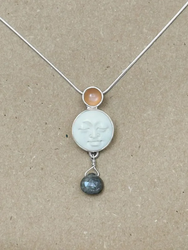 Sterling silver pendant necklace featuring a hand-carved bone moon face cabochon with a peach moonstone above and a faceted labradorite briolette drop below, on an 18" snake chain, displayed on kraft paper.