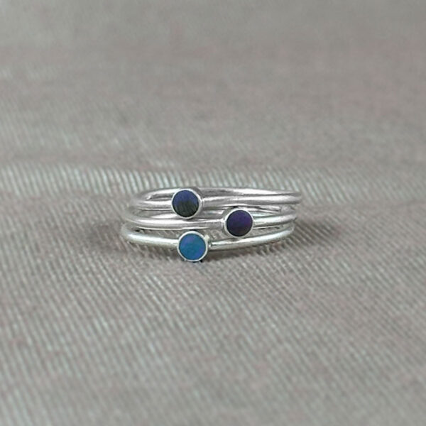 Three sterling silver stacking rings with hand-tinted clay dots in teal, dark blue, and gray, photographed on a linen-toned fabric background.