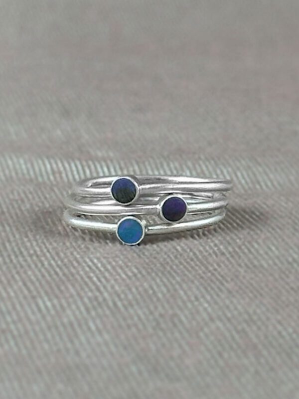 Three sterling silver stacking rings with hand-tinted clay dots in teal, dark blue, and gray, photographed on a linen-toned fabric background.
