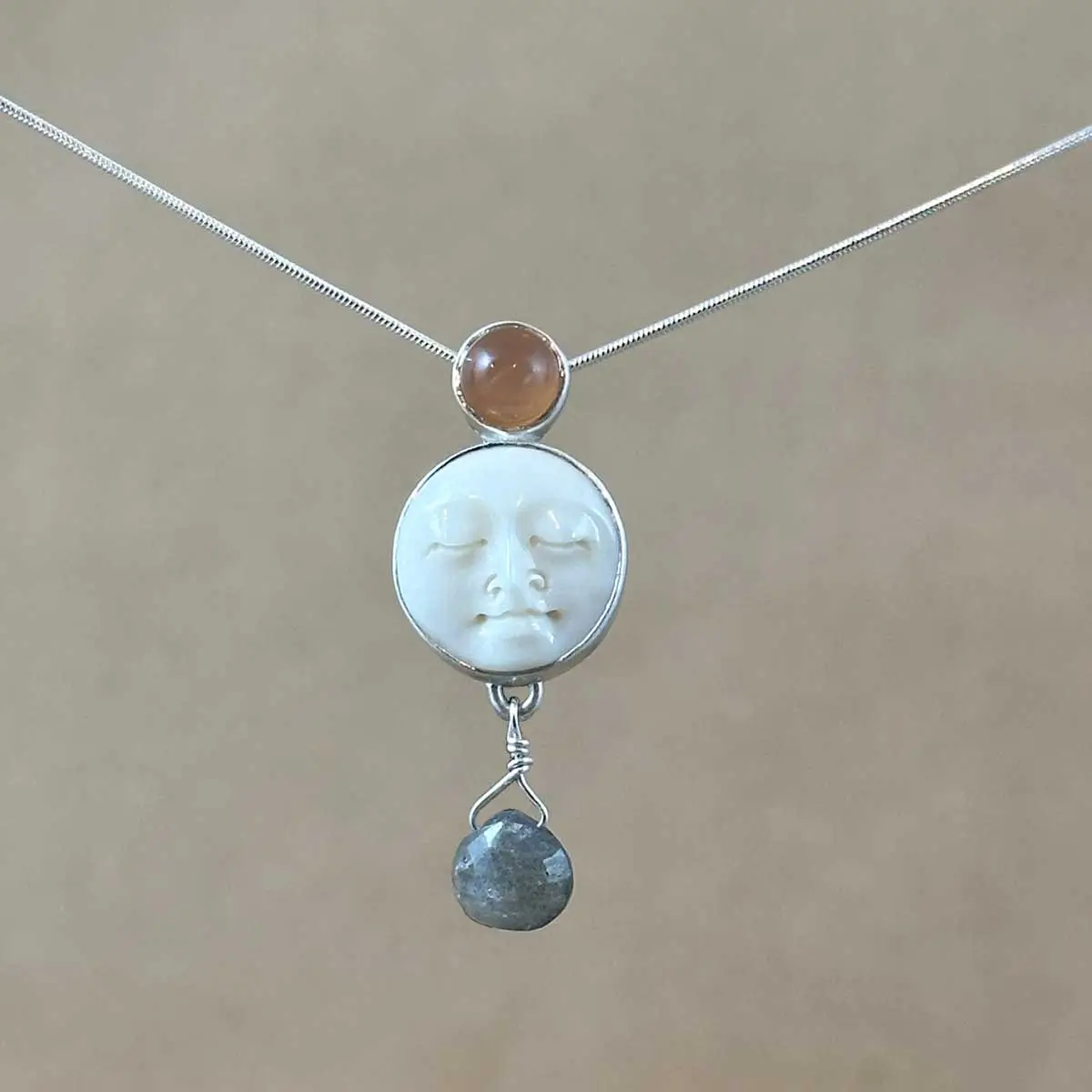 Sterling silver pendant necklace featuring a hand-carved bone moon face cabochon with a peach moonstone above and a faceted labradorite briolette drop below, on an 18" snake chain.