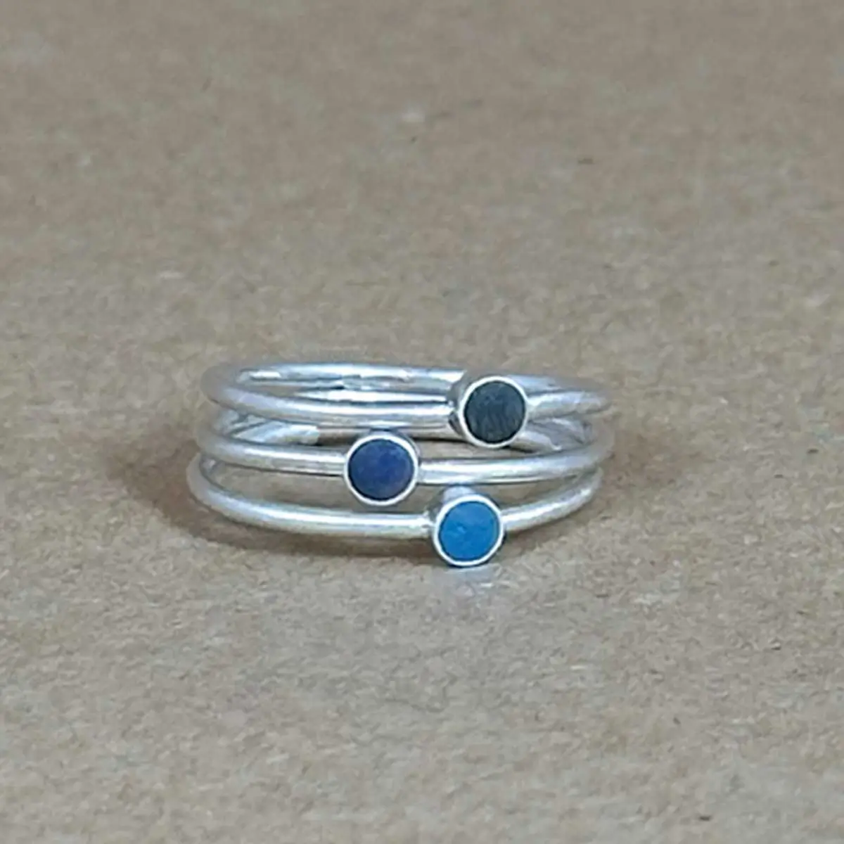 Three sterling silver stacking rings with hand-tinted clay dots in teal, dark blue, and gray, photographed on a linen-toned fabric background.